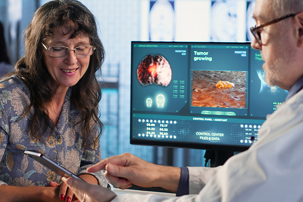 Female patient smiles as clinician shows information on a tablet, with medical monitors displaying brain images in the background.