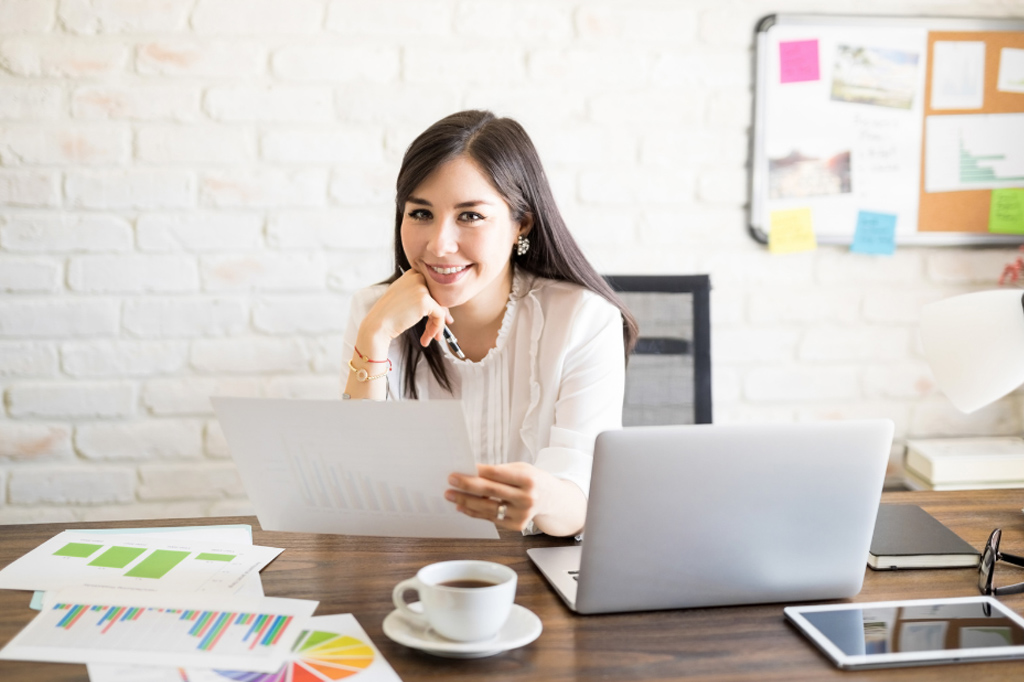 Businesswoman at a desk reviewing a financial report with charts, a laptop, and coffee point in hand.