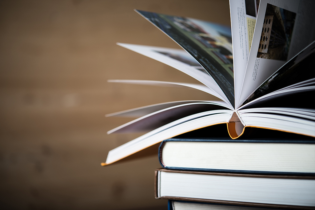 Stack of hardcover books with one open on top, pages fanned outward in a fan-like spread.