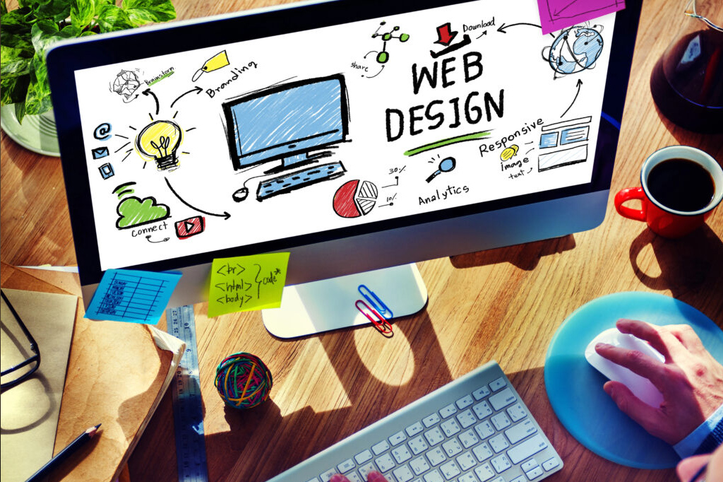 Desk workspace with a monitor showing a hand-drawn web design sketch, colorful sticky notes, keyboard and mouse on a blue pad, coffee mug nearby.