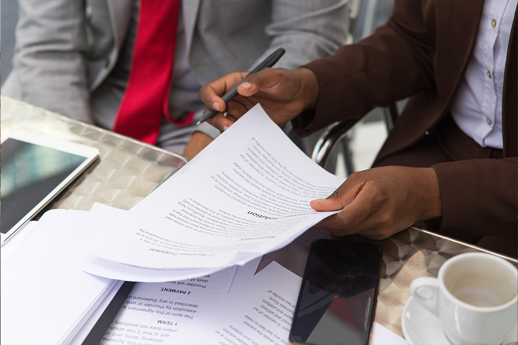 Two professionals review printed documents at a glass table, with a tablet, a smartphone, and a coffee cup nearby while one holds a pen over the papers.