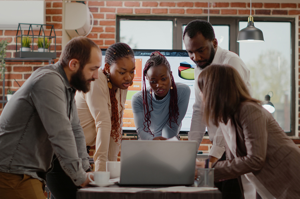 A diverse team leaning over a laptop on a table, collaborating in a modern brick-walled office as they review a presentation on screen behind them.