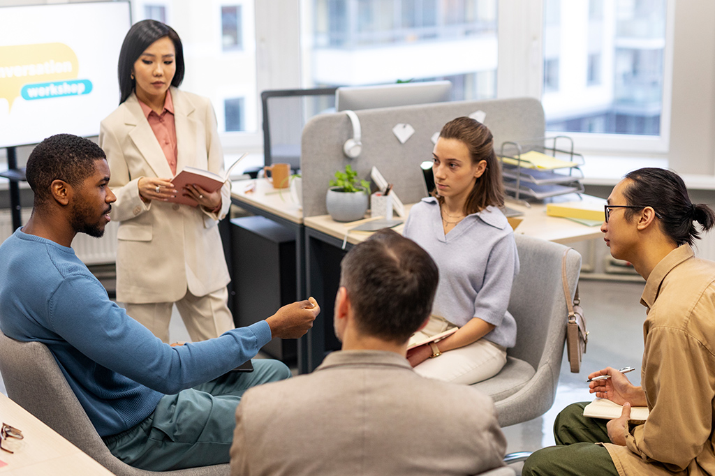 A diverse group of colleagues sits in a circle in a bright office, discussing during a workshop as one person presents with a notebook.