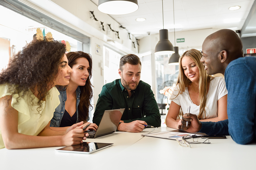 Five diverse coworkers seated around a bright office table, reviewing documents and a tablet together.