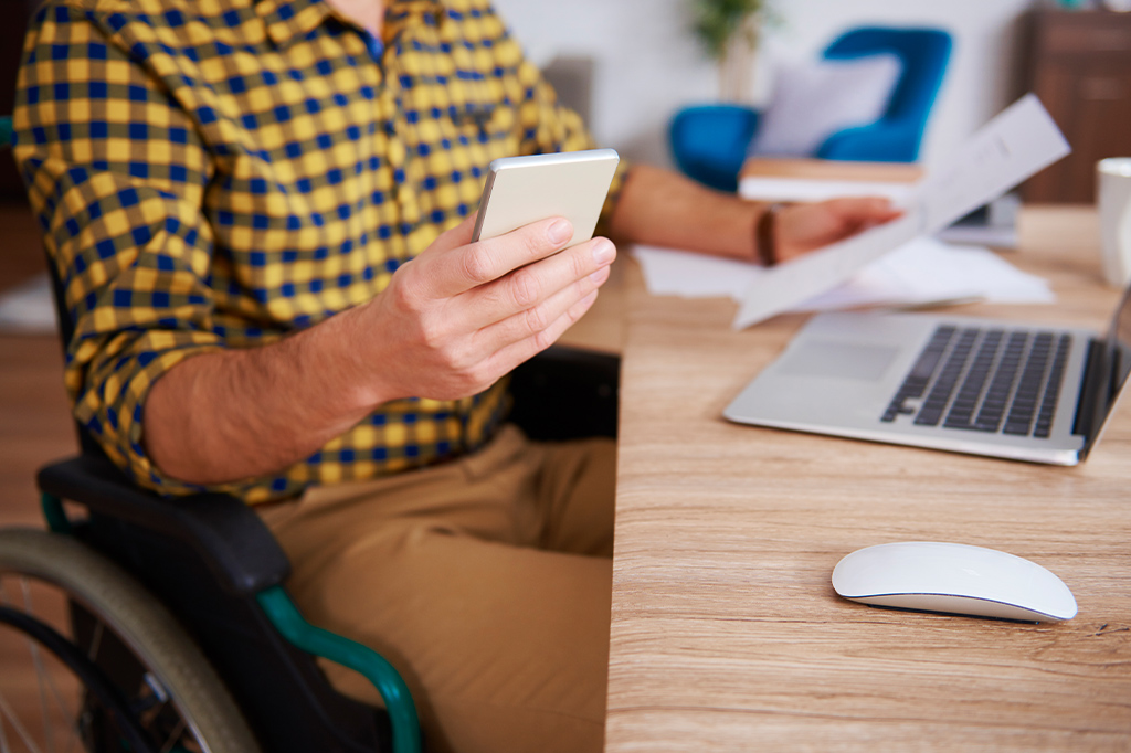 Person in a wheelchair at a wooden desk using a smartphone, with a laptop, papers, and a white mouse on the desk nearby.